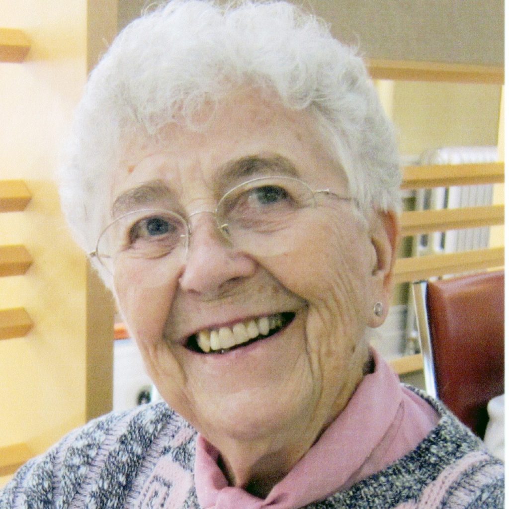A woman with short, white hair and round, wire glasses smiling for a picture wearing a pink scarf and a patterned blouse indoors.
