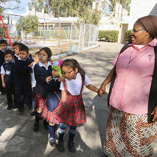 A teacher holding hands with a young girl in a school uniform. The young girl is a line leader of the line of her classmates headed inside from the playground.