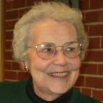 A woman smiling and looking to her left for a headshot indoors, with short grey hair, glasses, red earrings and a dark green sweater with a dark red brick background.