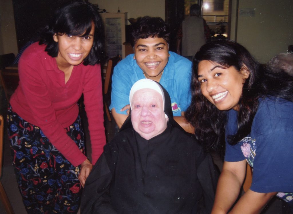 A nun sitting in a wheelchair smiling for a picture with three friends with dark hair indoors.