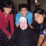 A nun sitting in a wheelchair smiling for a picture with three friends with dark hair indoors.