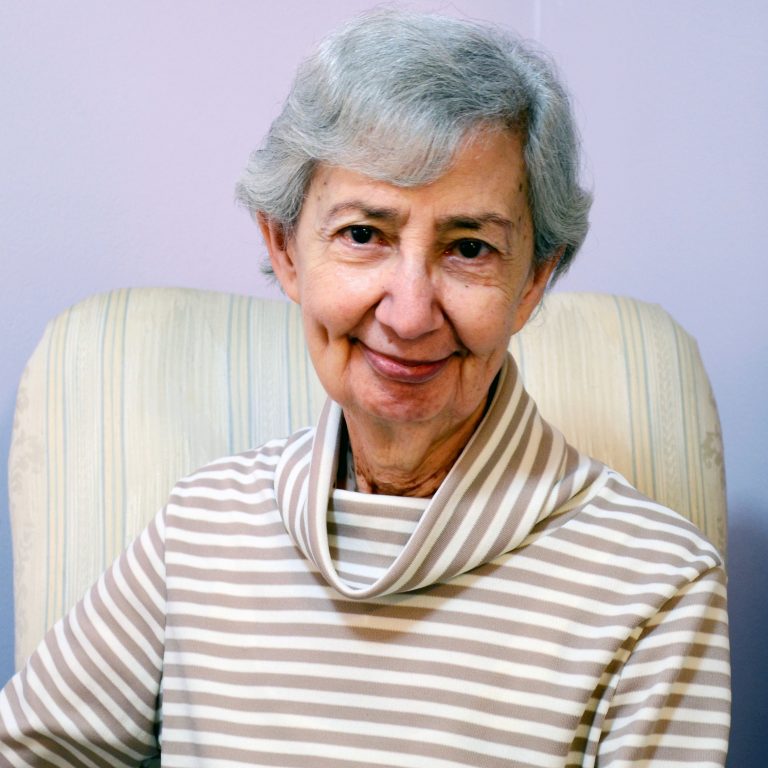 A woman with short, grey hair wearing a beige and white striped turtleneck sweater smiling while sitting in a white striped chair with a lilac background.