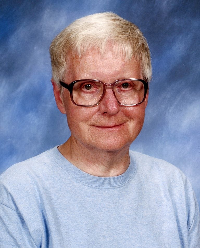A portrait headshot of a woman with short, white hair and glasses, wearing a baby blue sweater with a plain darker blue background.
