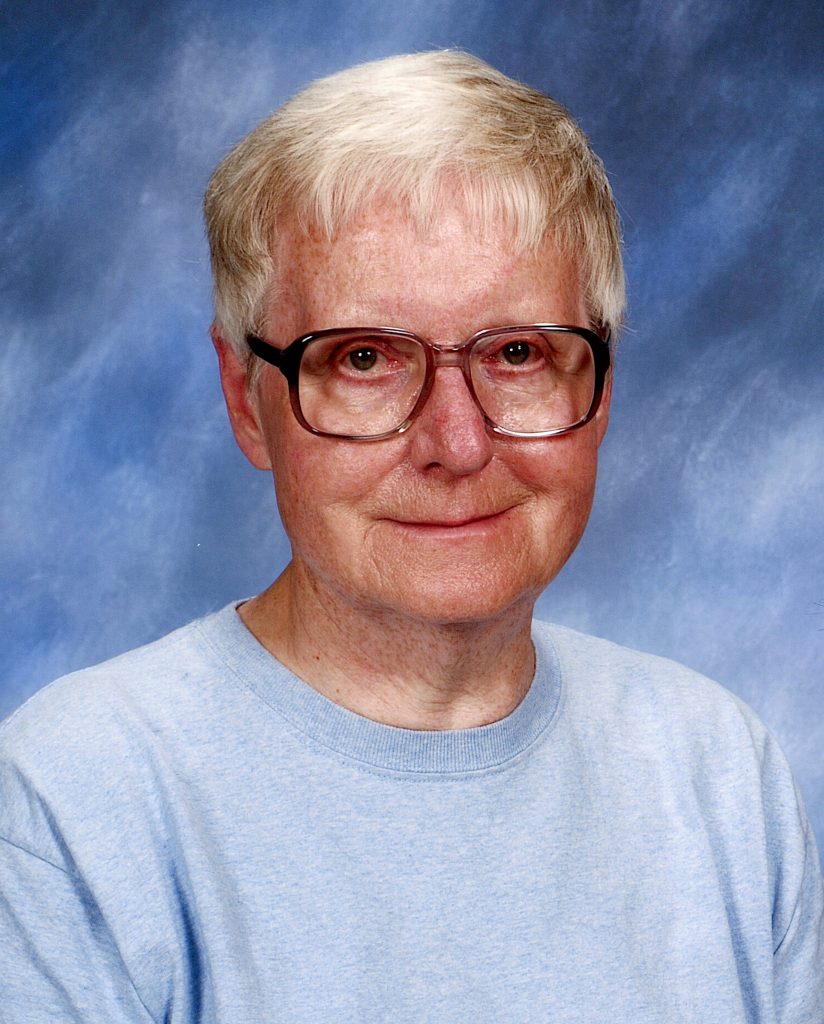 A portrait headshot of a woman with short, white hair and glasses, wearing a baby blue sweater with a plain darker blue background.