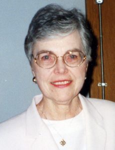 A woman with dark grey hair, and round wire glasses smiling for a picture indoors wearing a white shirt and light colored blouse, a necklace and small hoop earrings.