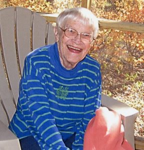A woman with short, grey hair, smiling for a picture while sitting in a lawn chair outdoors in autumn. She is wearing a dark blue striped sweater with a small green graphic on the front.