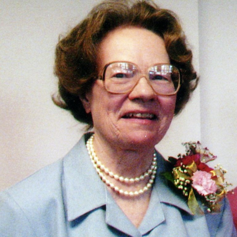 A woman with short brown hair and big glasses, wearing a powder blue blazer with two pearl strands and a pink boutonnière with a plain white background.