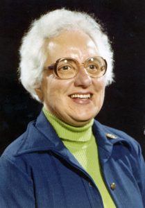 A woman smiling with short white hair, big brown framed glasses, a light green turtleneck, and a blue blazer with a black background.