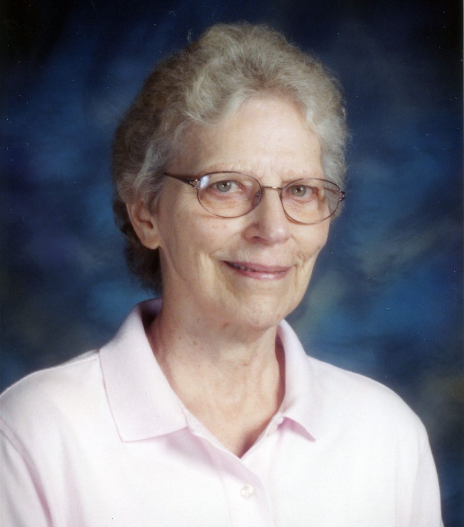 A portrait picture of a woman in a pale pink button up collared shirt with brownish grey hair and wire glasses with a dark blue background.