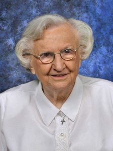 A woman with short grey hair and small wire glasses smiles for a portrait picture wearing a white collared shirt with lace detailing and a cross necklace with a blue background.