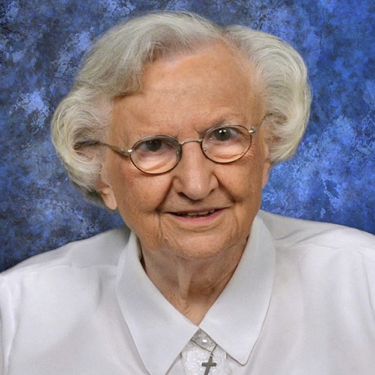 A woman with short grey hair and small wire glasses smiles for a portrait picture wearing a white collared shirt with lace detailing and a cross necklace with a blue background.