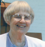 A woman with short light colored hair and big glasses smiling for a picture wearing a lavender shirt, a white cardigan and a white flower boutonnière.