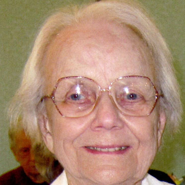 A woman with short white hair and big glasses smiling for a headshot picture in a room wearing a white collared button down shirt.