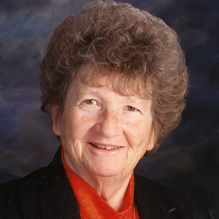 A portrait headshot of a woman with short brown hair smiling in a red blouse, with a black blazer, in front of a dark grey background.