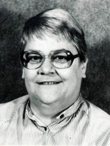 A black and white headshot of a woman with short hair, and dark framed glasses, wearing a collared blouse, and smiling for a picture with a dark background.
