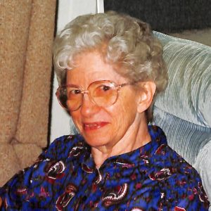 A woman with short curly grey hair, wearing big glasses, and a dark blue patterned collared shirt smiling for a picture while sitting on a plush grey velvet chair indoors.