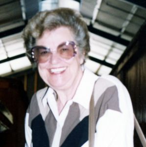 A woman with short, dark, grey hair, and big round sunglasses, wearing a white collared shirt with black and grey geometrical patterns, smiling for a picture.