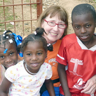 A woman with red-framed glasses smiling brightly for a picture with three young children outdoors.