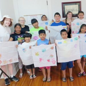A class of young kids smiling brightly while displaying their tye-dye art projects with their three teachers indoors.