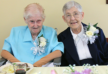 Two women with short grey hair, wearing dress clothes and white rose and ribbon boutonnières smiling brightly together while sitting and enjoying a meal at a table.
