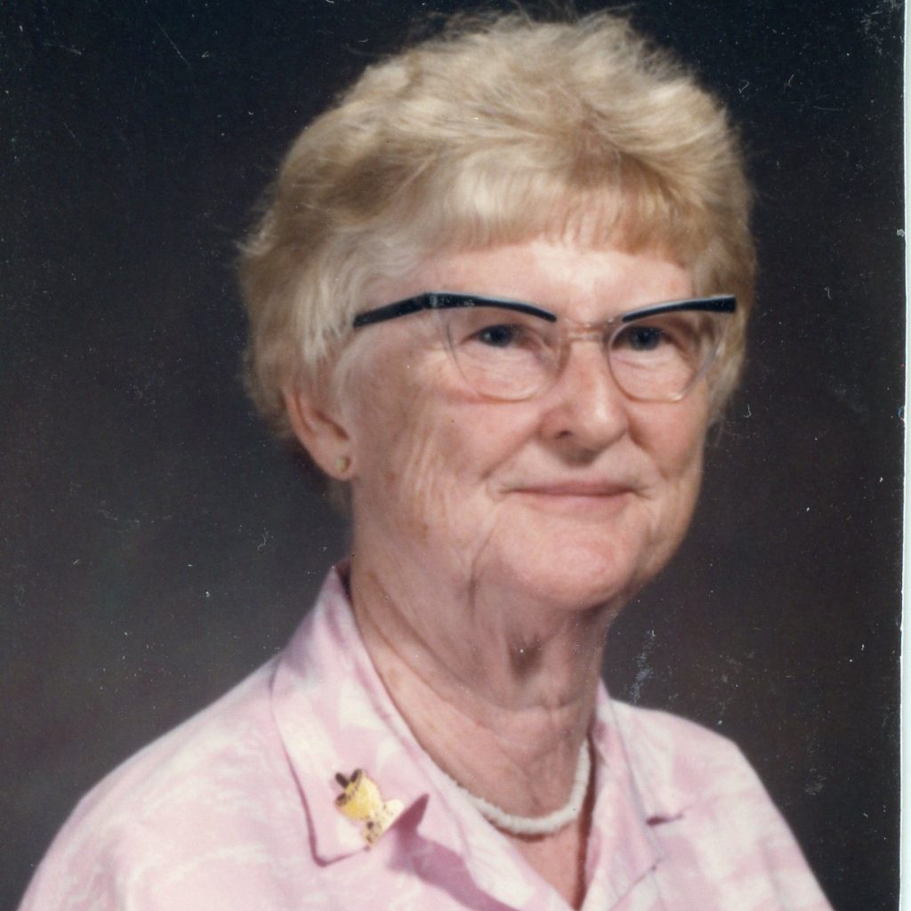 A woman smiling for a headshot picture, with light colored short hair, and glasses, wearing a pearl necklace and a light pink collared blouse with a plain dark grey background.