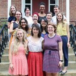 Thirteen women smiling together for a group picture on the front stairs of a red brick building outdoors.