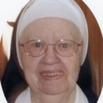 A nun wearing round wire glasses smiling for a headshot picture.