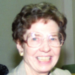 A woman with short brown hair, earrings and big glasses smiling brightly for a headshot picture.