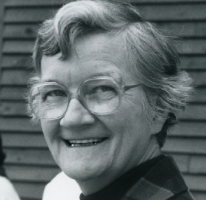 A black and white headshot of a woman smiling with short grey hair, and big glasses.