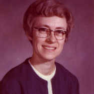 A woman smiling for a portrait picture with short brown hair, glasses, a white shirt and a dark colored cardigan with a dark grey background.
