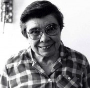 A black and white headshot of a woman smiling with short dark hair, and big glasses, wearing a plaid collared shirt.