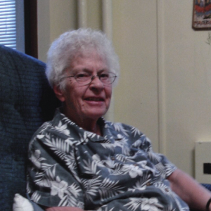 A woman sitting in a chair indoors, smiling, and looking to her right for a picture with short grey hair, round wire glasses, a grey patterned shirt.