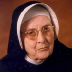 A nun wearing glasses smiles for a headshot picture with a dark brown background.