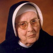 A nun wearing glasses smiles for a headshot picture with a dark brown background.