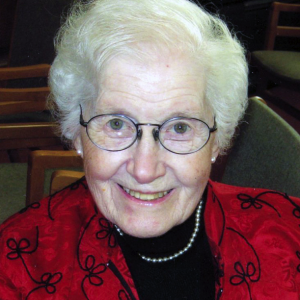 A woman smiling for a headshot picture wearing round, black wire glasses, a black shirt, a red patterned blouse and a pearl necklace.