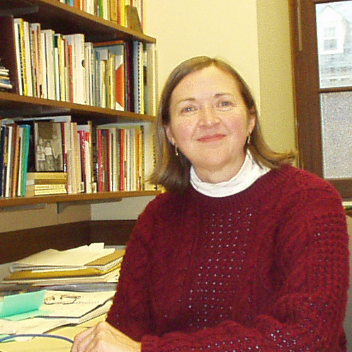 A woman with shoulder length brown hair, wearing a white turtleneck, a red sweater, and dangly earrings smiling for a picture in an office space at a desk with shelving holding books and butter yellow walls in the background.