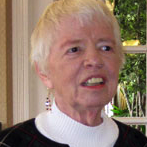 A woman smiling for a headshot picture, with short white hair, wearing dangly earrings, a white turtleneck and a dark cardigan.