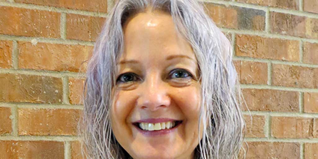 A woman with shoulder-length grey hair wearing a grey and white sweater with a red brick background.