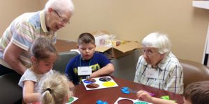 Grandparents and three grandchildren play a board game at a wooden table indoors.
