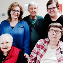 Five women, two sitting and three standing together for a group picture indoors.