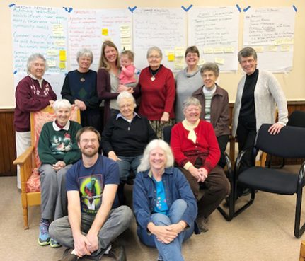 A group picture in a class room, two individuals are sitting on the floor, three are sitting in chairs, and eight standing behind in an inverted triangle shape.