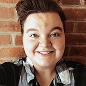 A woman with short brown hair smiling brightly for a headshot picture wearing a black and white plaid collared shirt with a red brick background.