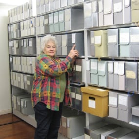 A woman with grey hair wearing a colorful plaid shirt smiling while grabbing a file box on a large file shelf covering the whole wall.