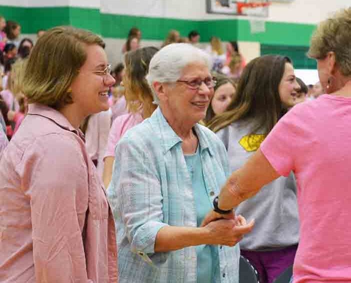 Three women connect in a crowd, shaking hands and greeting each other with warm smiles.