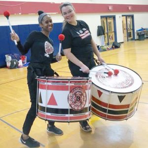 Two women playing drums for the marching band in a school gym.