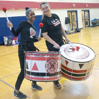 Two women playing drums for the marching band in a school gym.