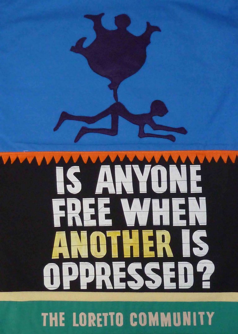 Handmade banner with a graphic of a fat person standing on top of a thin person laying down. Text reads "Is anyone free when another is oppressed? The Loretto Community"
