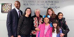 Three young girls, three women, and one man standing together in front of a hand-painted wall sign stating: Mary Helen Sandoval Café "Let us remember: one book, one pen, one child, and one teacher can change the world." - Malala Yousafzai