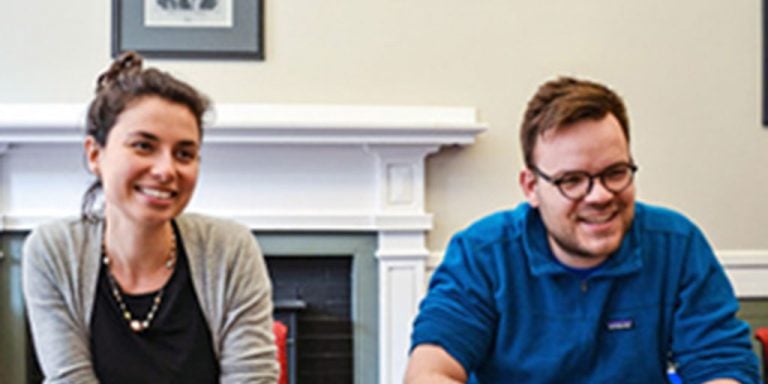 A man and a woman sitting next to each other smiling and laughing candidly at a dark wooden table in a home in front of a white fire place.