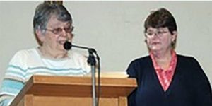 Two women, both with short brown hair and glasses standing together while speaking into a microphone on a wooden podium in front of a plain white wall.